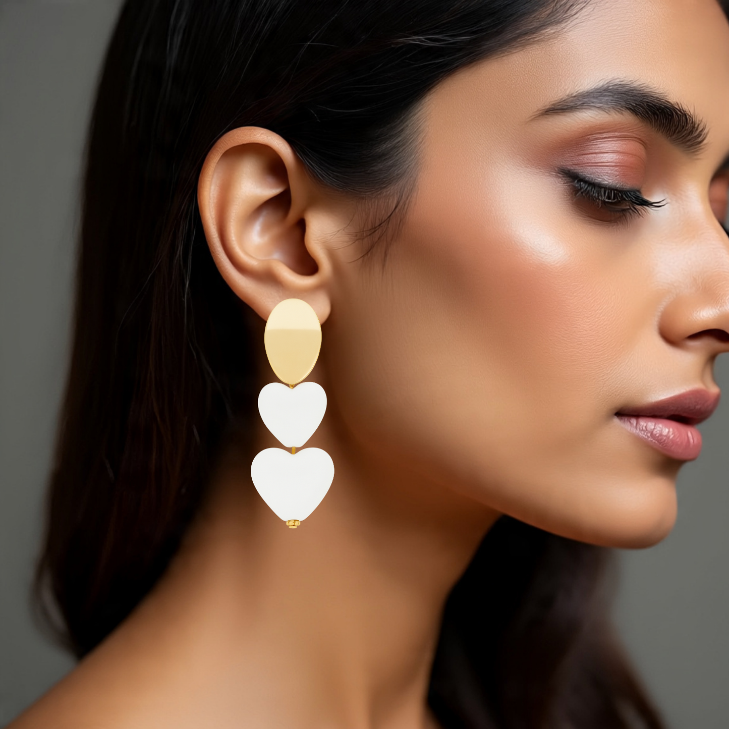 Close-up of a woman wearing gold and white heart-shaped earrings against a neutral background