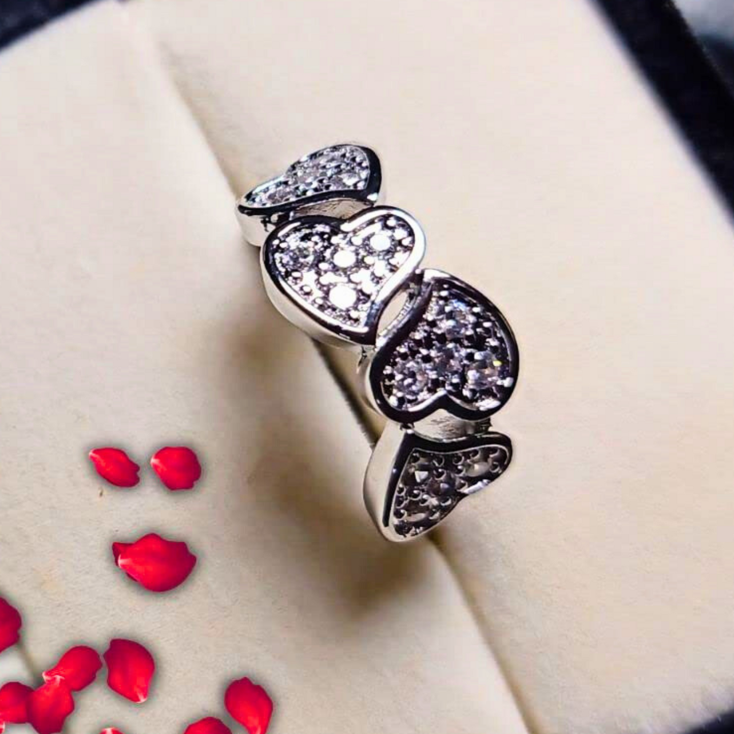 Silver ring with heart-shaped gemstones on a beige surface with red rose petals.