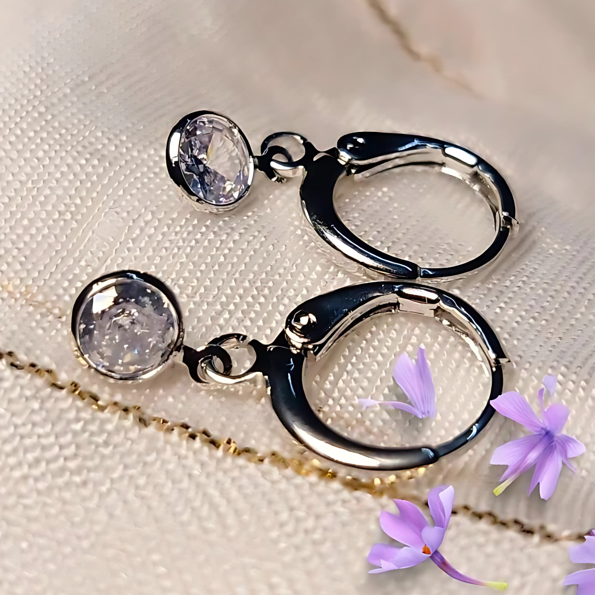 Silver hoop earrings with clear gemstones on a textured beige surface with purple flowers.