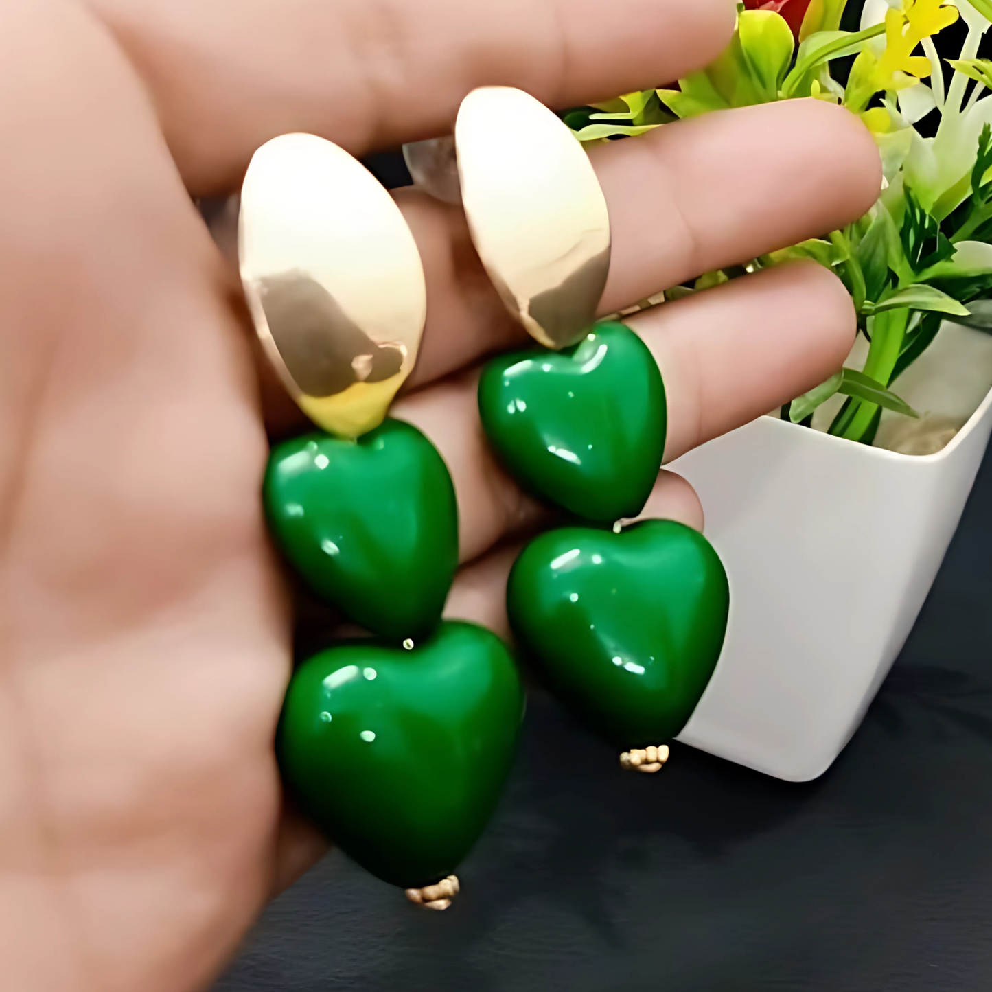 Green heart-shaped earrings with gold accents held by a hand, with a plant in the background.