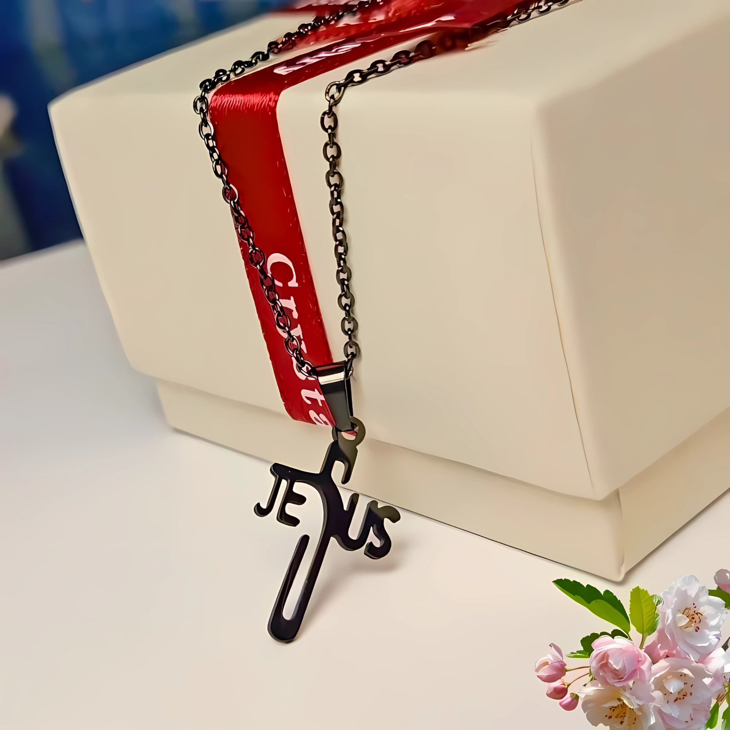 Necklace with 'JESUS' pendant on a red ribbon with 'Cristmas' text, placed on a white surface with flowers.