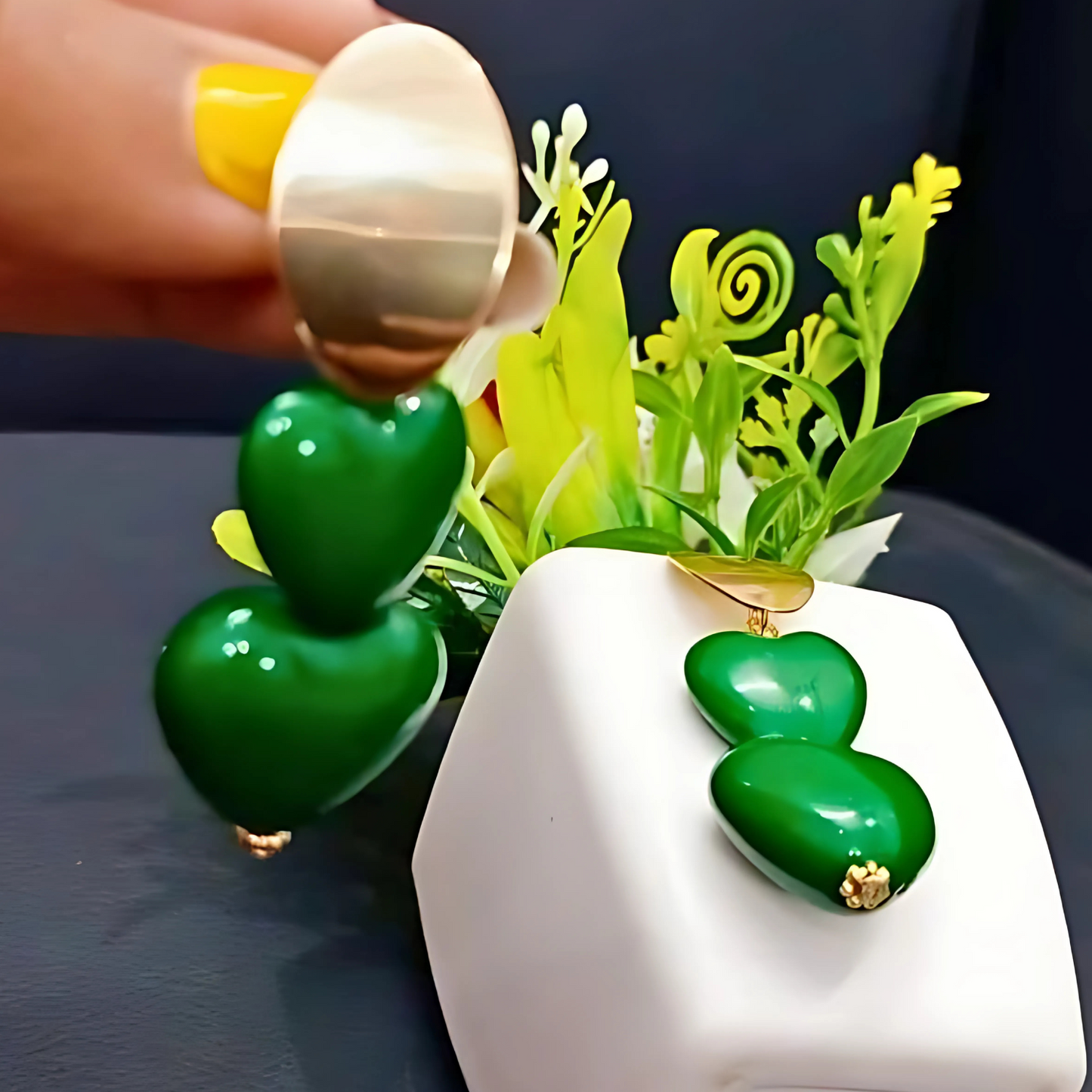 Green heart-shaped earrings with gold accents displayed on a white stand with decorative plants.