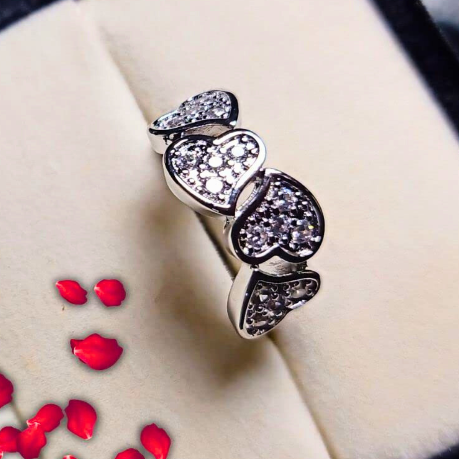 Silver ring with heart-shaped gemstones on a beige surface with red rose petals.