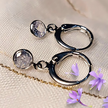 Silver hoop earrings with clear gemstones on a textured beige surface with purple flowers.