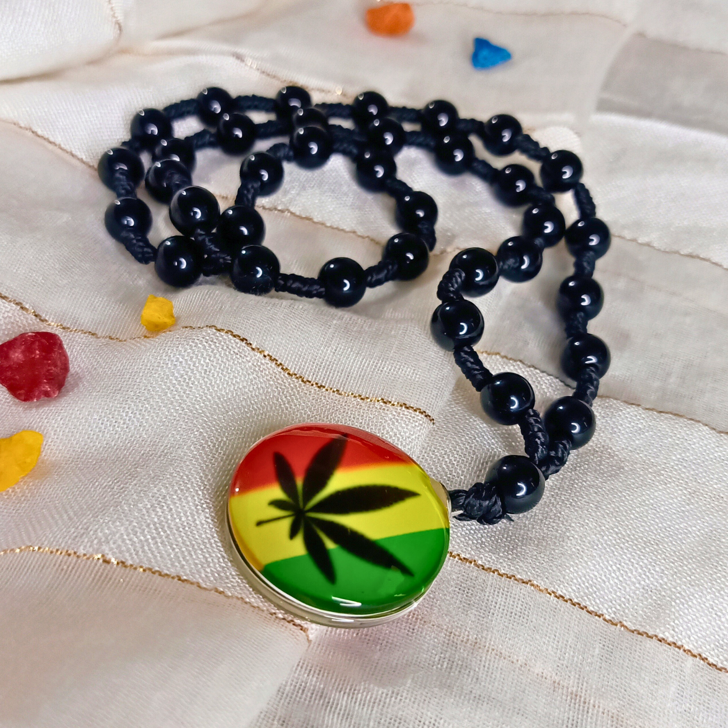 Black beaded necklace with a colorful pendant featuring a cannabis leaf design on a textured fabric background.