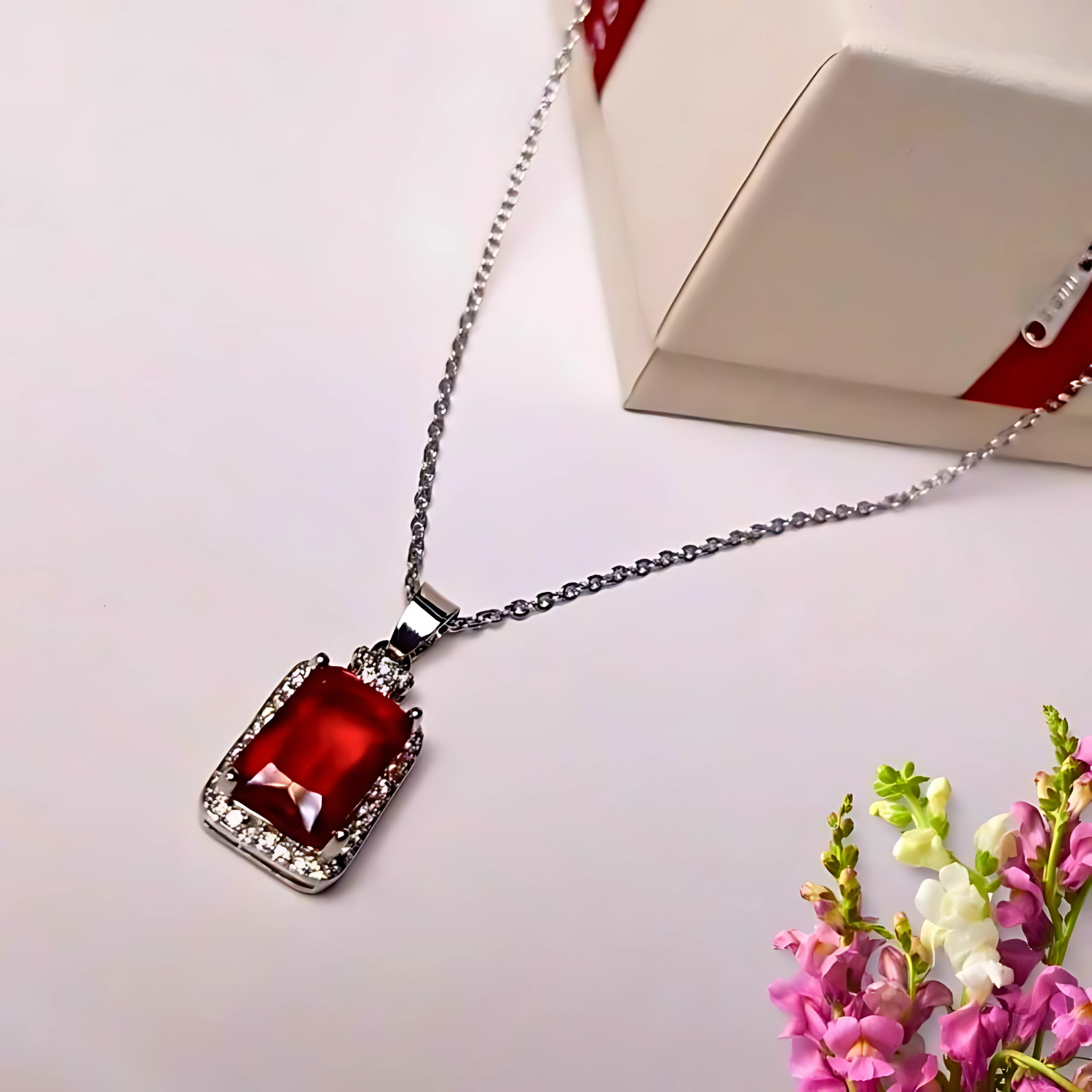 Necklace with a red gemstone pendant on a white surface with flowers and a box in the background