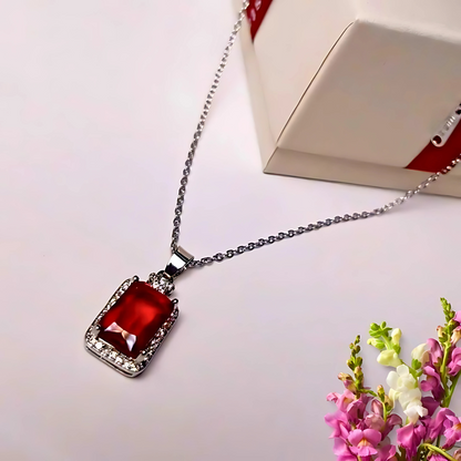Necklace with a red gemstone pendant on a white surface with flowers and a box in the background